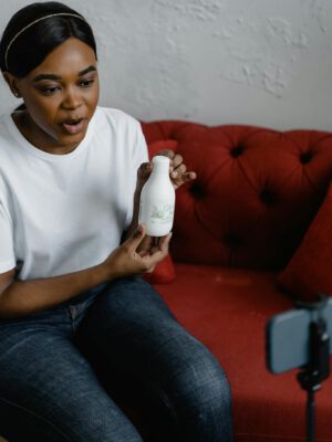 African American woman presenting a cosmetic product while vlogging on a red sofa.
