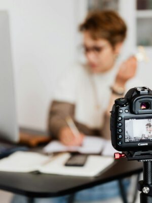 Unrecognizable blurred lady working at table in modern office against photo camera placed on tripod